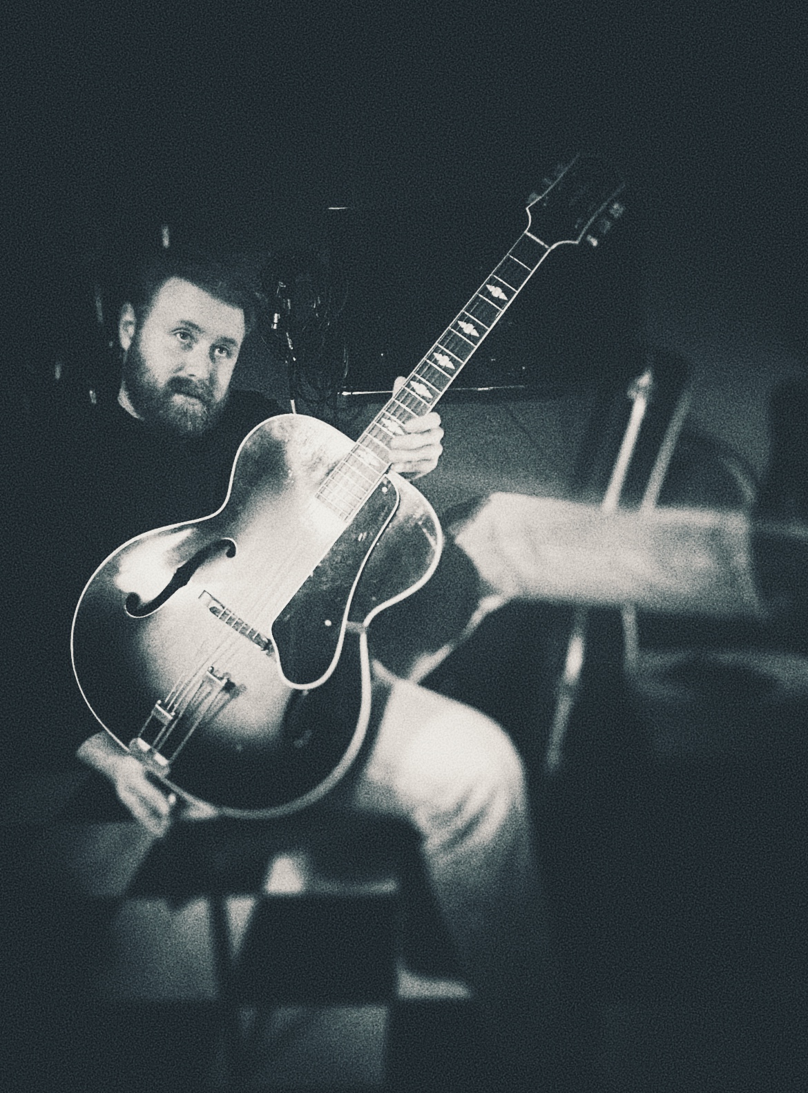 A guitarist performing with a vintage archtop jazz guitar, captured in black and white with dramatic lighting and a focused, concentrated expression.